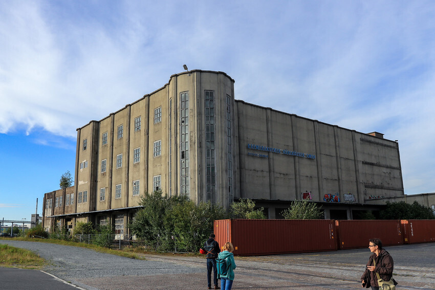 Zu sehen ist ehemaliger Hafenschuppen, davor stehen Container. Es handelt sich um die letzte freie Entwicklungsfläche der Überseestadt.